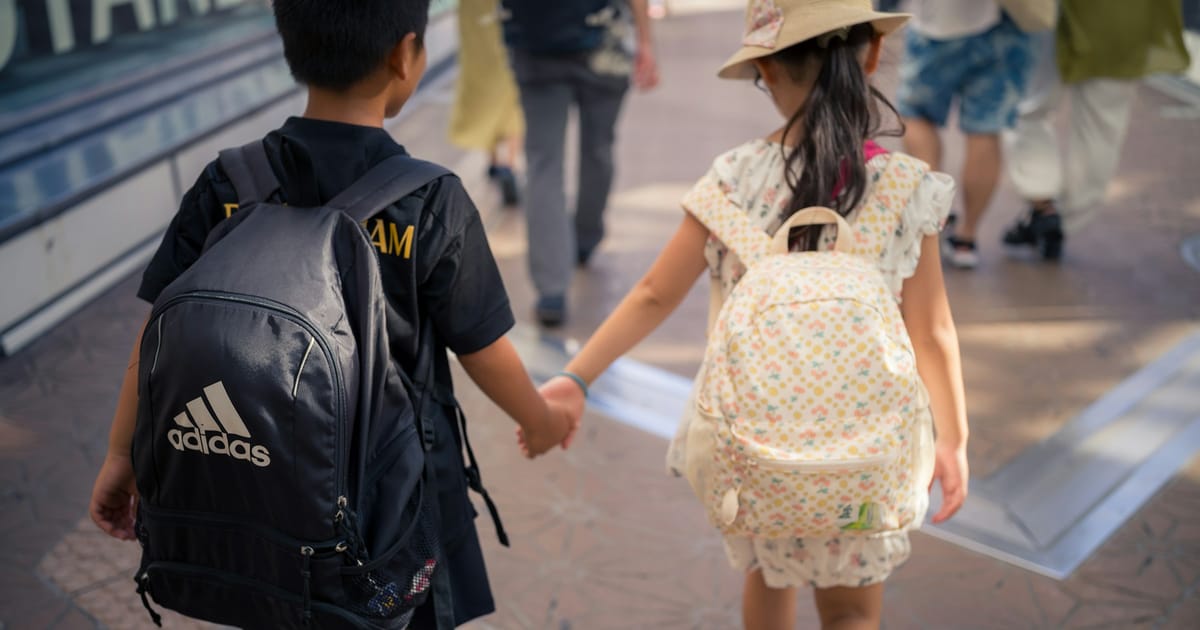 Dos niños con mochilas escolares caminan de la mano por una vereda, vistos de espaldas.