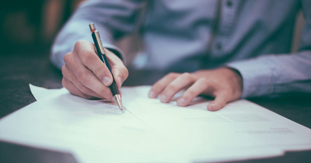 Una persona escribe con un bolígrafo sobre una hoja en un escritorio.