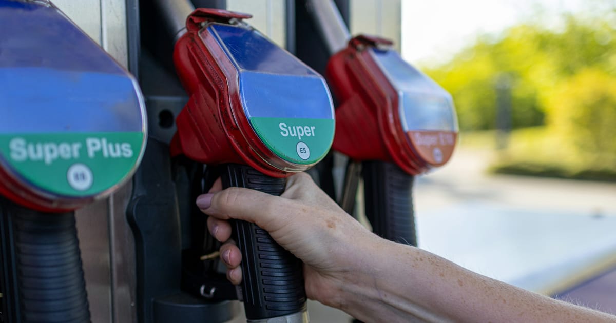 Una mano toma la manguera de un surtidor de gasolina, junto a otras mangueras de colores.